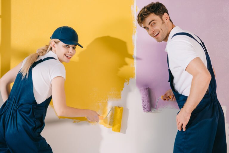 two cheerful painters looking at camera while painting wall in yellow and pink